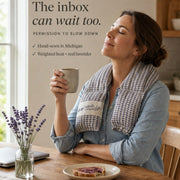 A woman sits at a wooden table in a warm kitchen setting, holding a mug with a knitted lavender-themed neck warmer draped around her shoulders. Text on the left reads, 'The inbox can wait too.