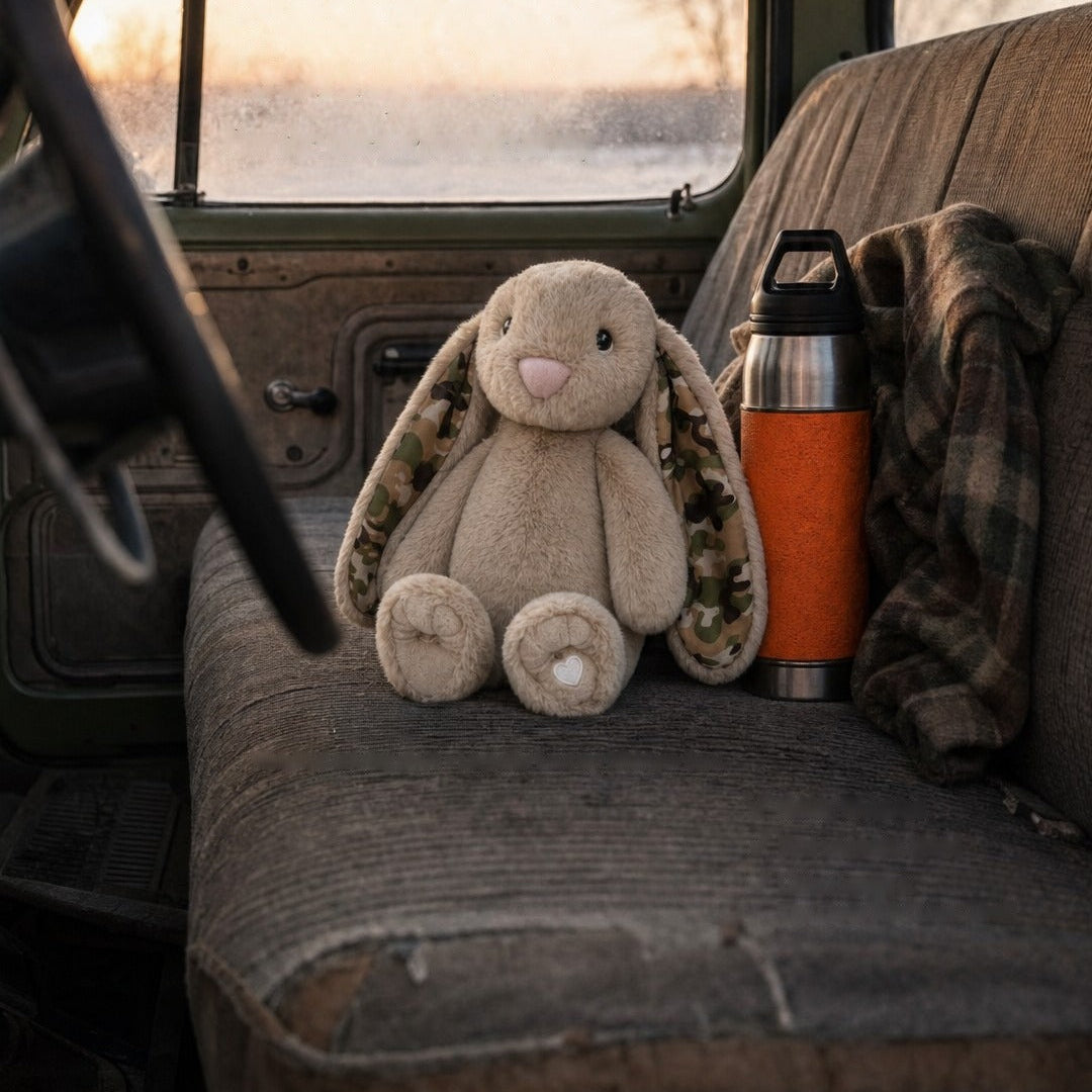 A beige plush bunny toy with camouflage-patterned inner ears sits on the back seat of a worn vehicle. An orange insulated bottle and a plaid blanket are beside it, with a window and snowy outdoor scene in the background.
